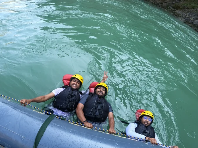 Guests body-surfing the rope line during a river rafting trip in Rishikesh — life jackets and helmets on, calm pool between rapids