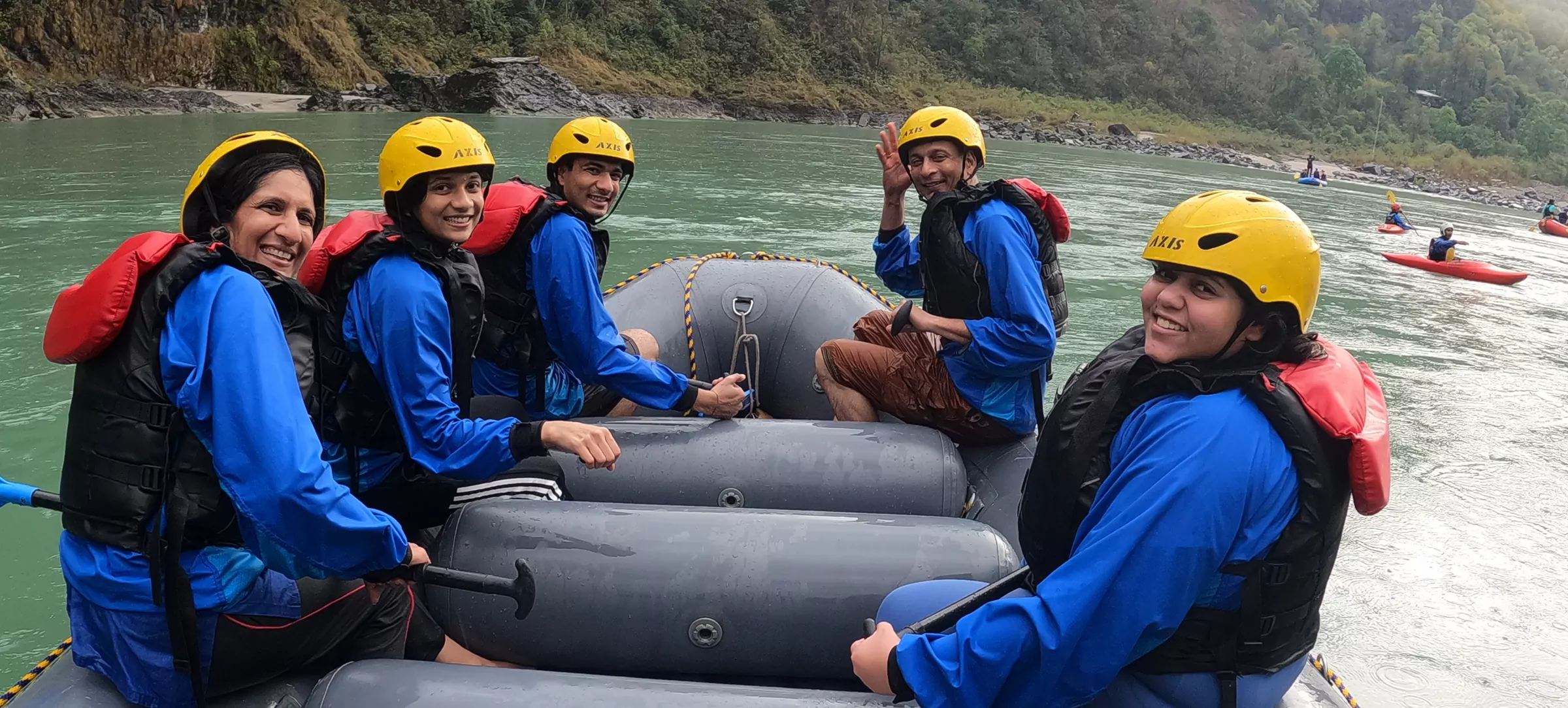 Smiling group on a raft mid-Ganga during whitewater rafting in Rishikesh — yellow helmets, blue thermal jackets, safety kayaker in the background