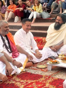 Priests and guests seated on the steps at Shatrughan Ghat before the Ganga Aarti, brass puja thali and marigold lamp in front of them, Rishikesh